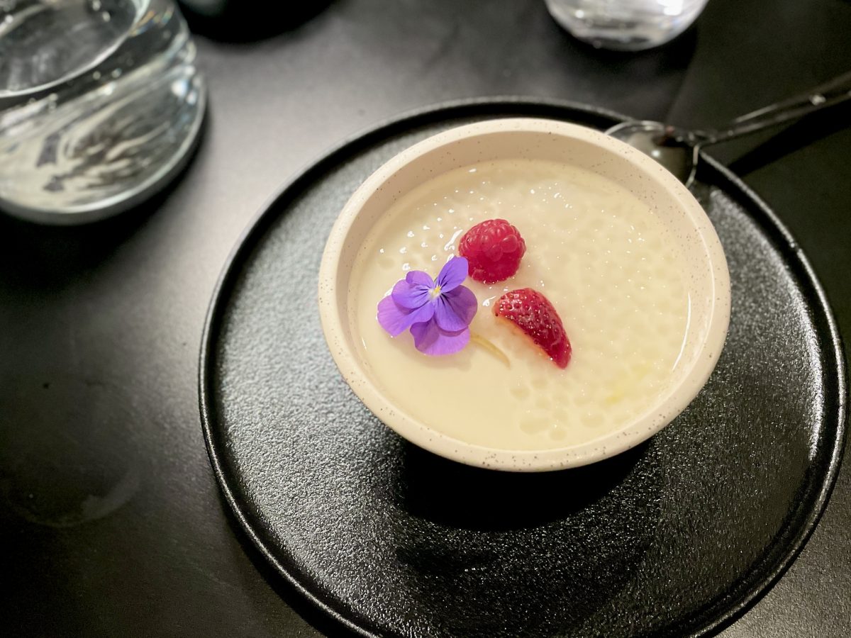 A white bowl with creamy dessert, fruit and a purple flower on a black table.
