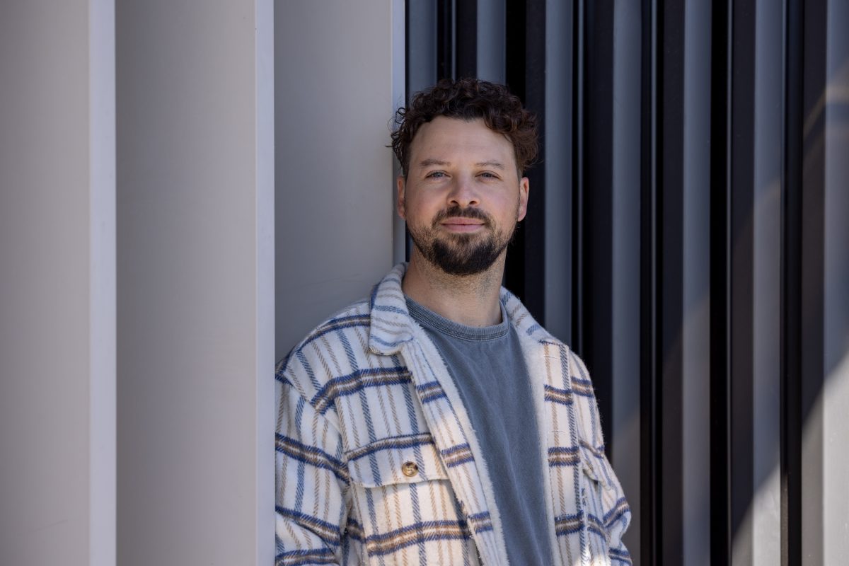 A bearded man in an open checked shirt stands in front of a wall with vertical panelling.