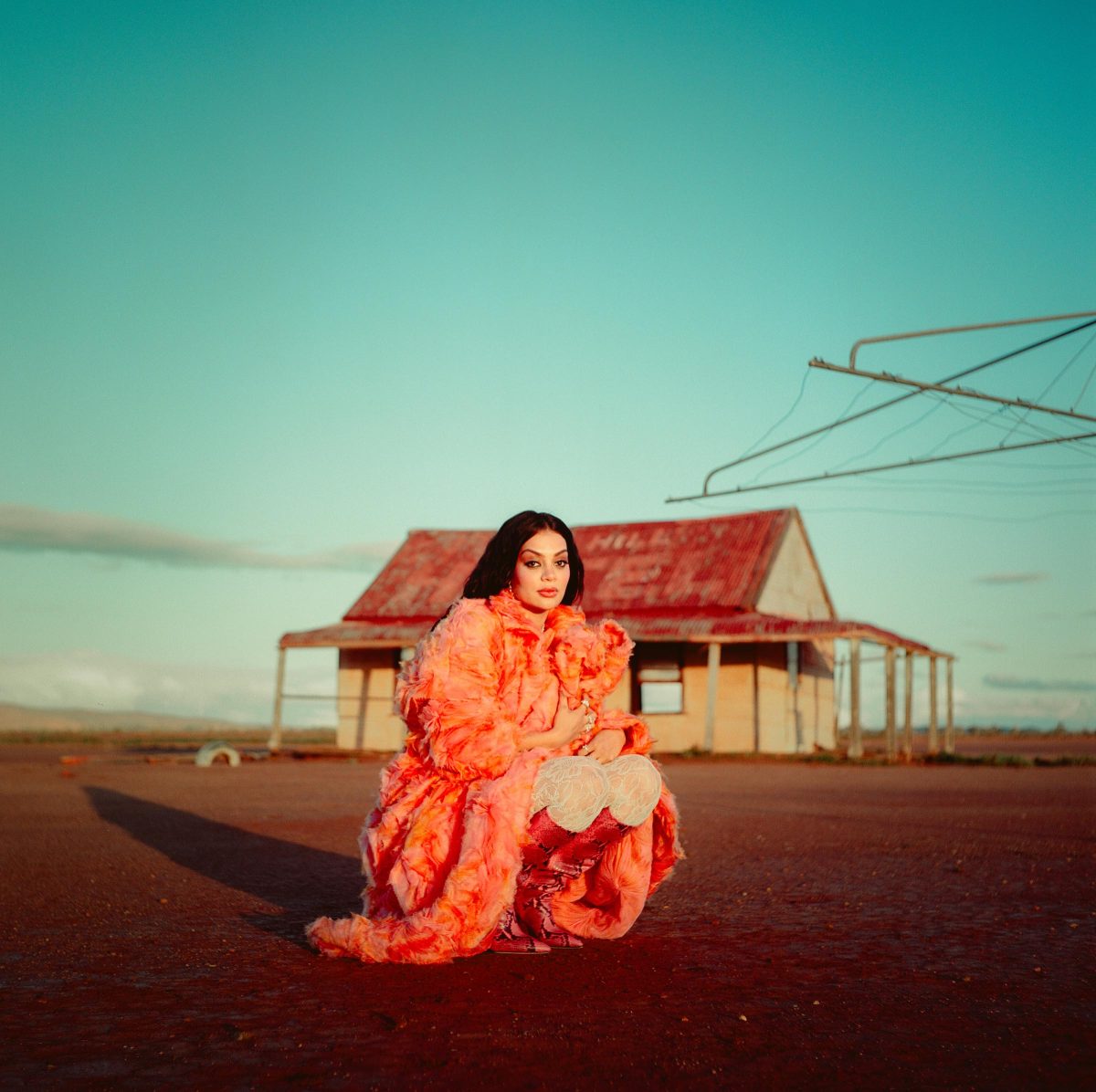 Thelma Plum crouching on dirt land in-front of a house wearing an orange jacket