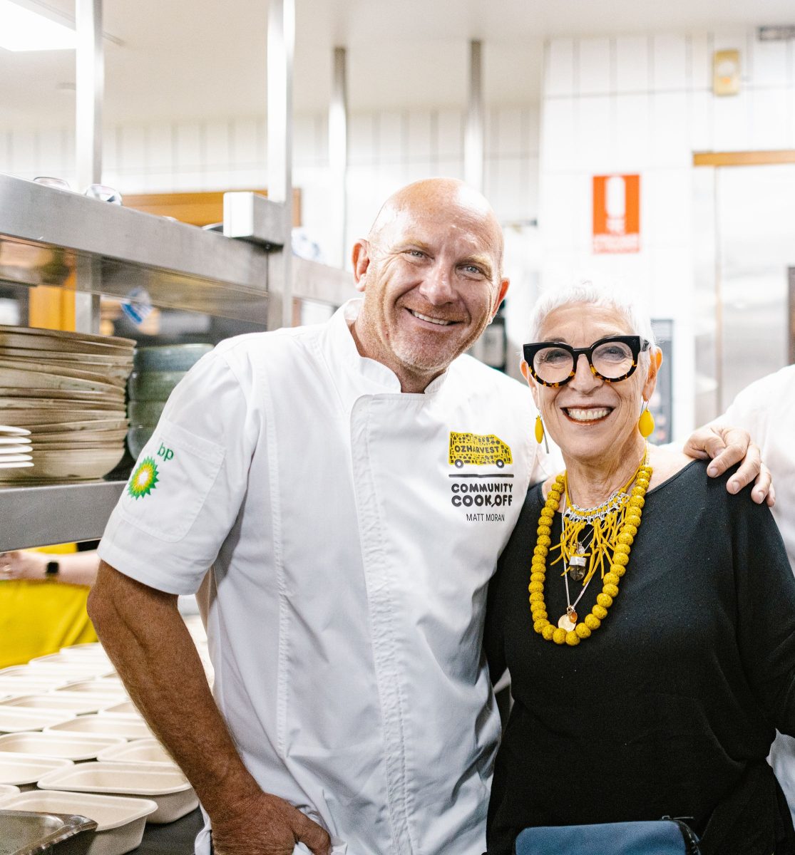 Chef Matt Moran and OzHarvest founder Ronni Kahn AO stand arm in arm in a kitchen.