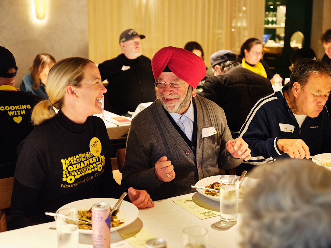 A woman in black and yellow OzHarvest shirt dines next to an older man wearing a red turban.