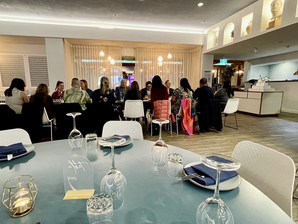 A table laid with wineglasses in the foreground with a long table of diners in the background.