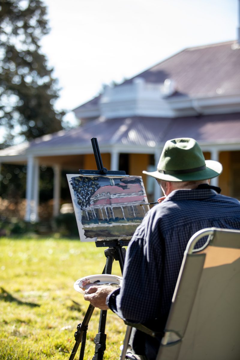 A plein-air (or 'open air') artist sits in front of an easel, creating an impression painting of a historic homestead