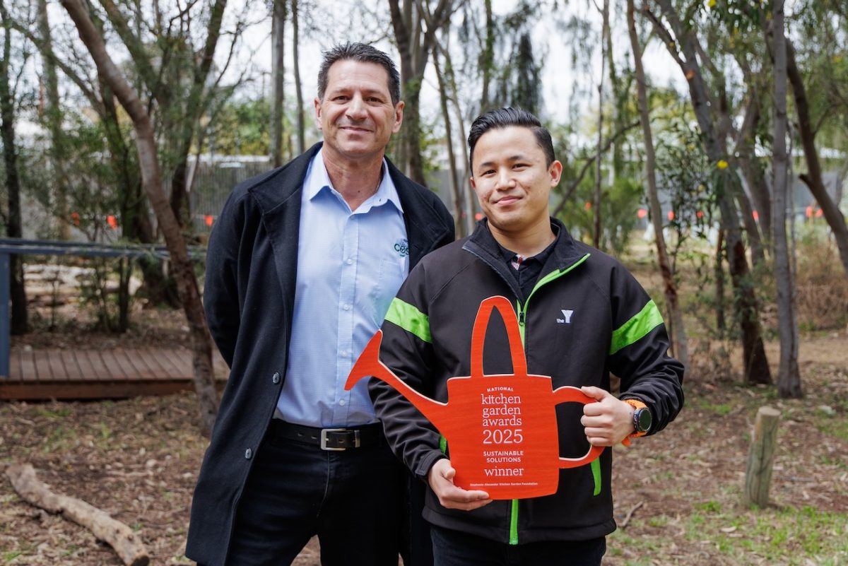 Two men stand in a garden, one holds a watering can-shaped award.