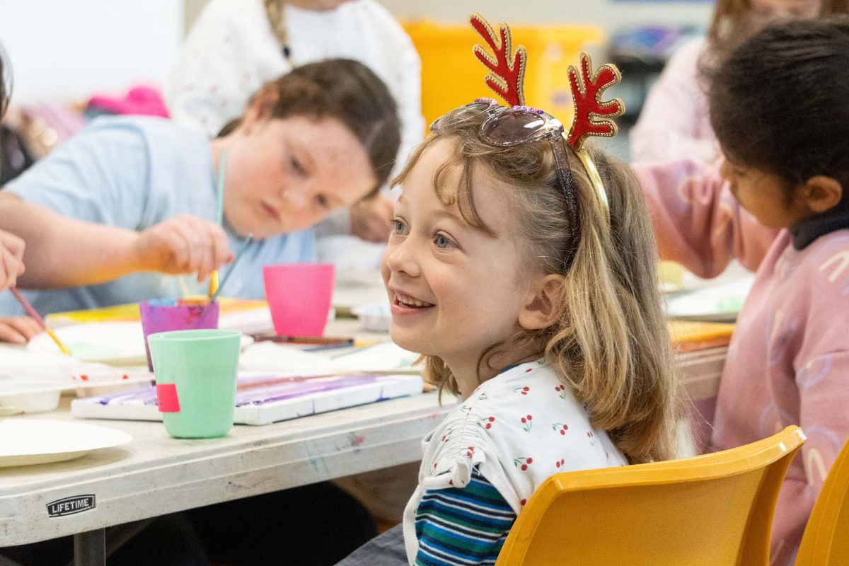 a child wearing reindeer antlers sitting at a table with paints and a canvas