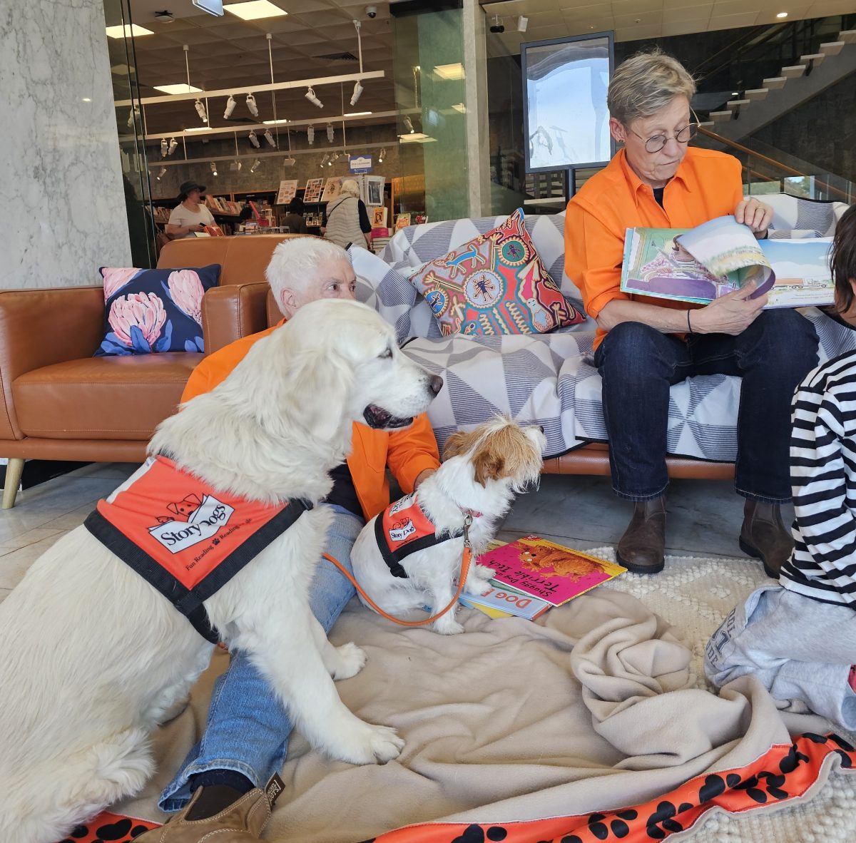 Story Dog volunteers dressed in bright orange uniforms read from a story book, while two dogs wearing matching vests appear to listen intently.