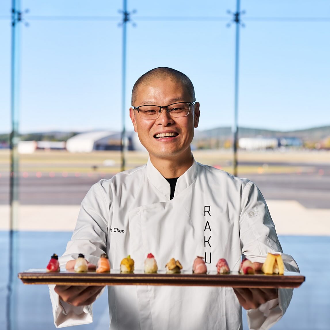 A Chef wearing white Raku branded jacket holds a long plate with different sushi.