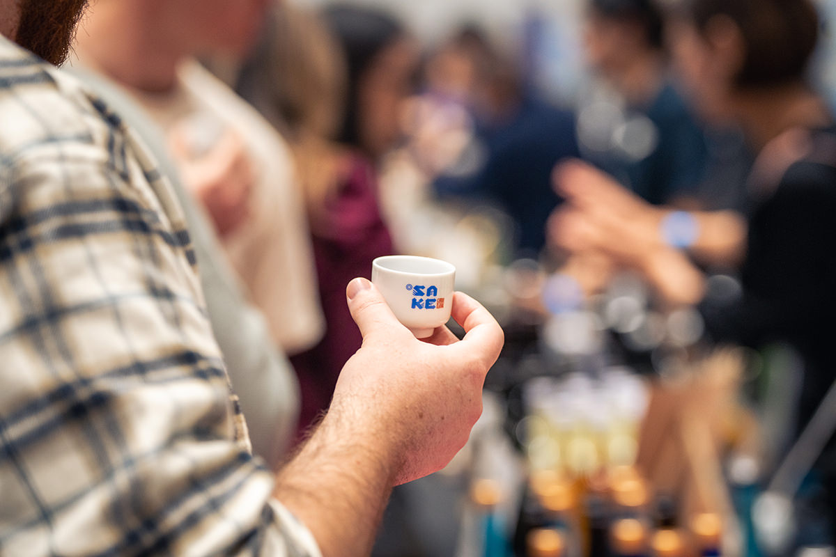 A hand holding a sake cup branded with the festival's logo.
