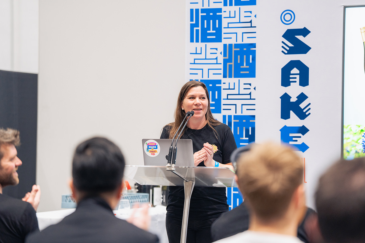 A woman stands at a podium in front of a blue and white sign reading Sake.