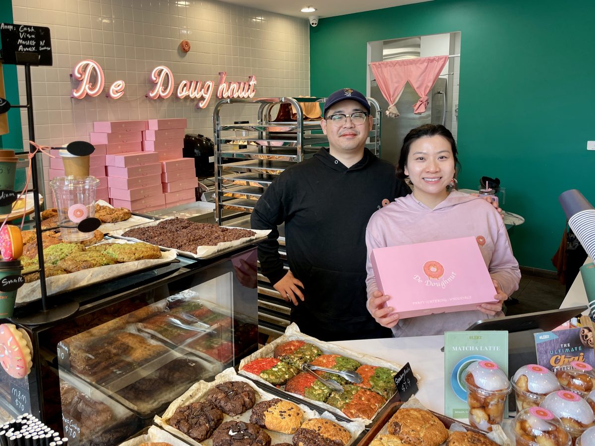 A man and woman stand behind a counter filled with cookies. She is wearing pink and holding a pink de doughnut branded box.