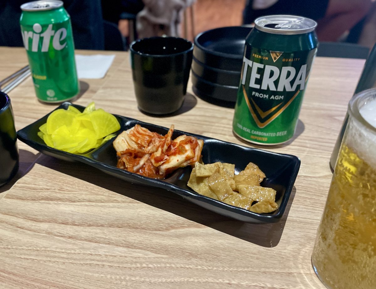 A small black dish separated into three parts filled with a yellow radish pickle, red kimchi and tan-brown fish cakes. A can of Terra beer is in the background.