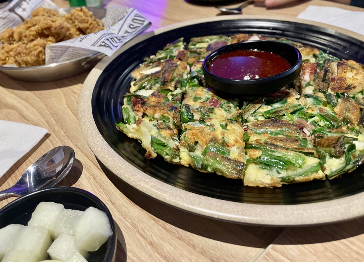 A savoury pancake with bowl of sauce in the centre. In the foreground, white pickled radishes and a plate of fried chicken in the back.