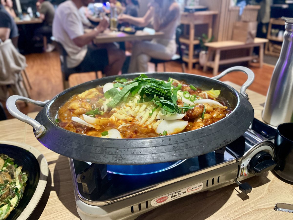 A wide metal dish on a hotplate in a restaurant. The dish contains ingredients covered in a red sauce, with cheese and spring onion on top.