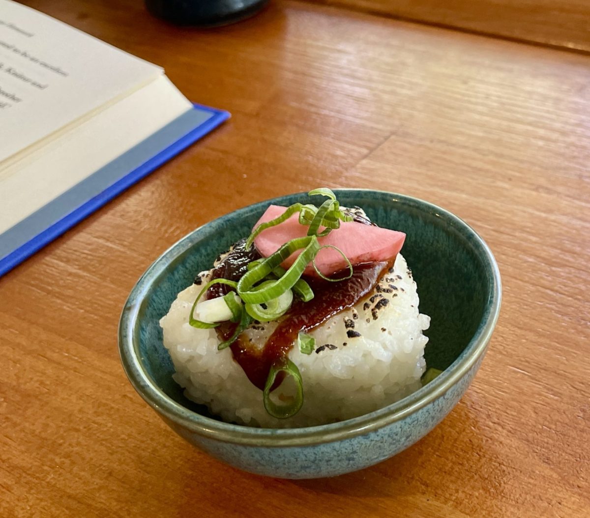 A small round dish with a triangle shaped rice ball, topped with a reddish sauce, spring onion and a slice of pink pickle.