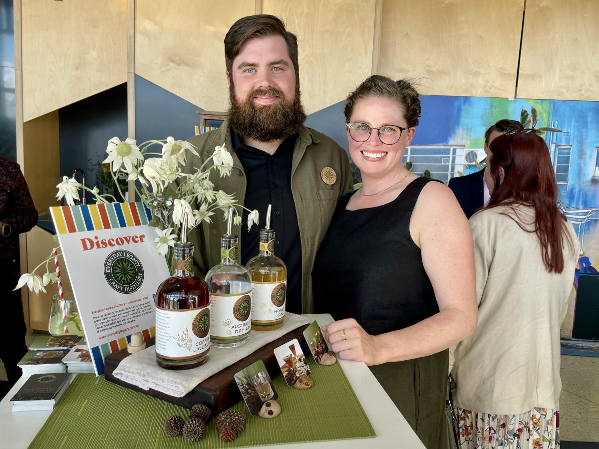 A bearded man and woman in glasses stand behind a display with three bottles of spirits.