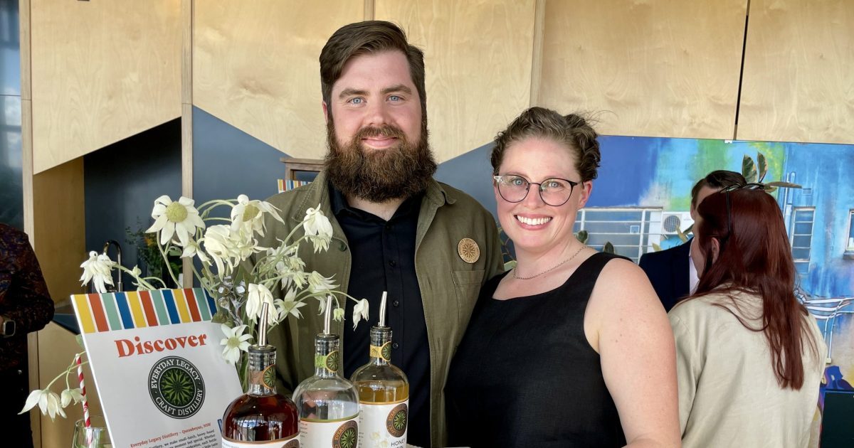 A bearded man and woman in glasses stand behind a display with three bottles of spirits.