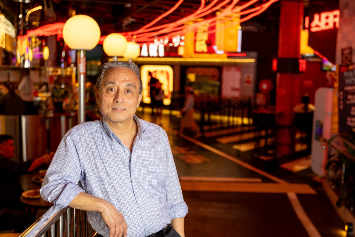 Older Chinese man leans against railing with red lights in background.