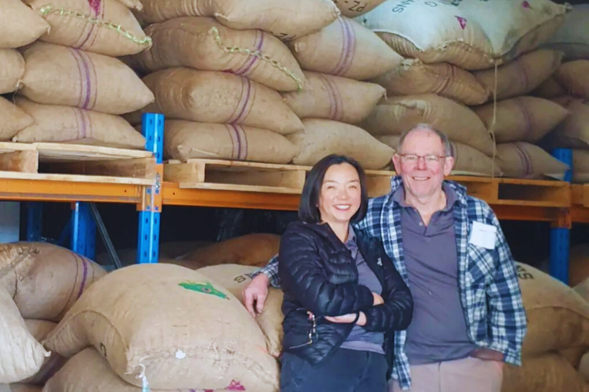 Man and woman stands in front of warehouse of cocao beans.