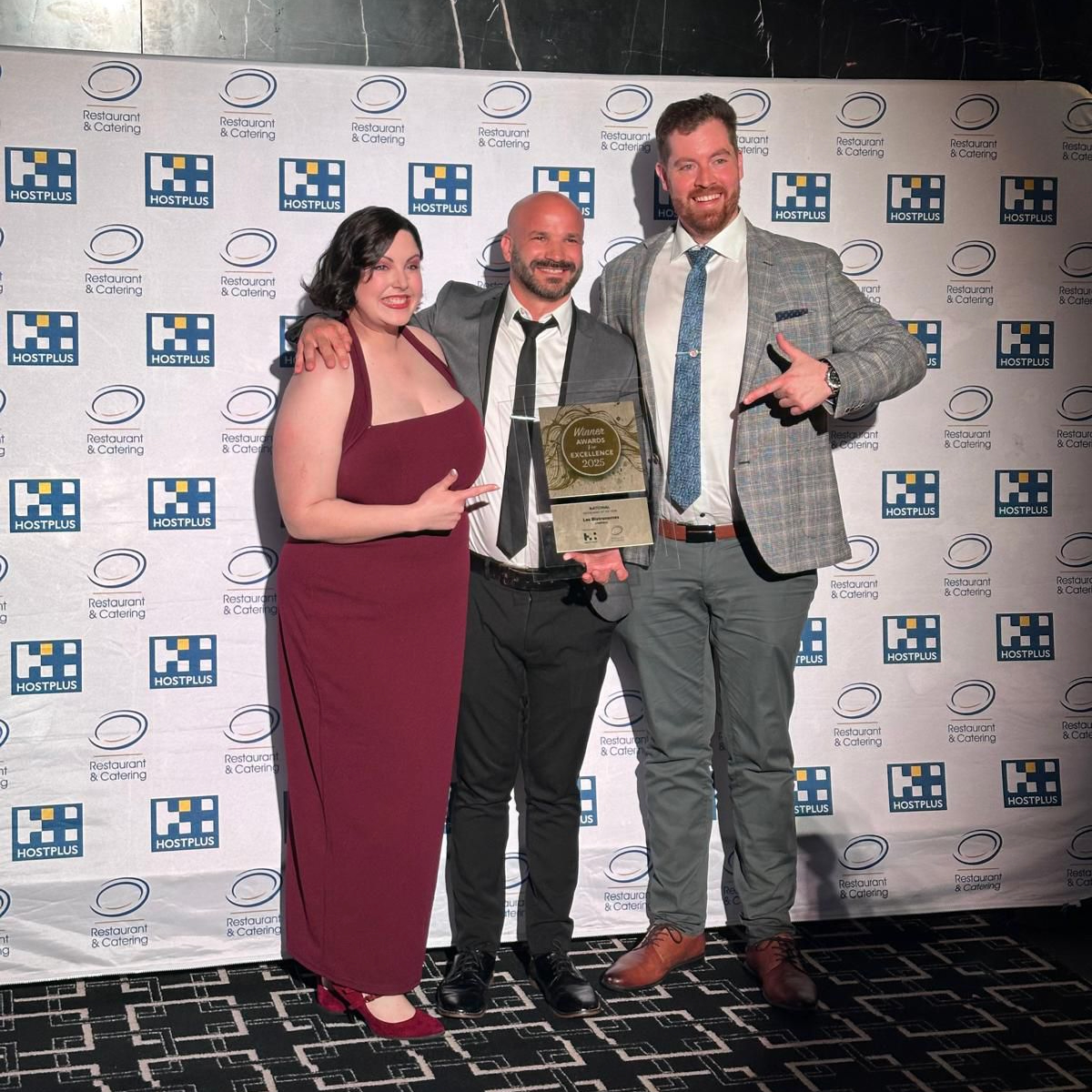 Three people smile in front of a branded R&CA backdrop. The person in the centre is holding an award and the other two are pointing at it and smiling.
