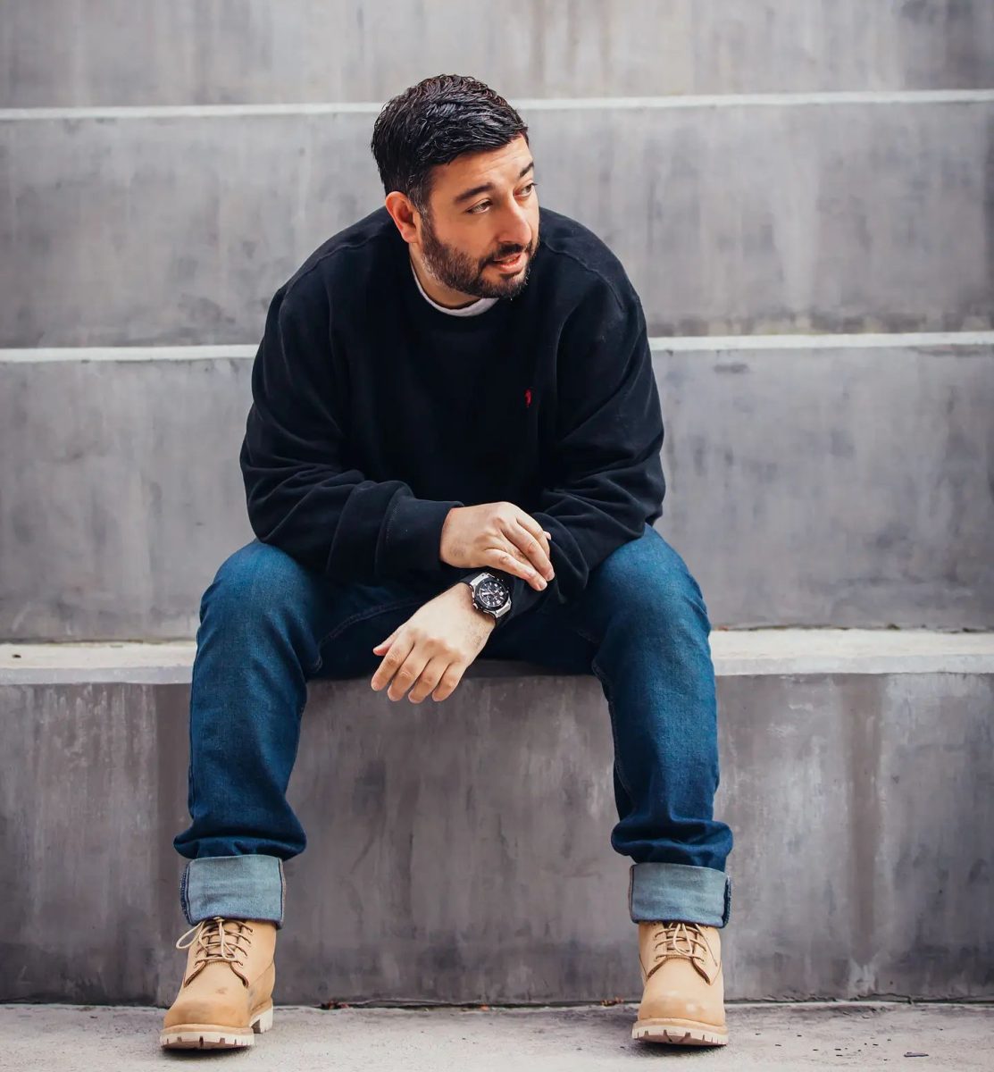 Man in black jumper and jeans sits on concrete steps.
