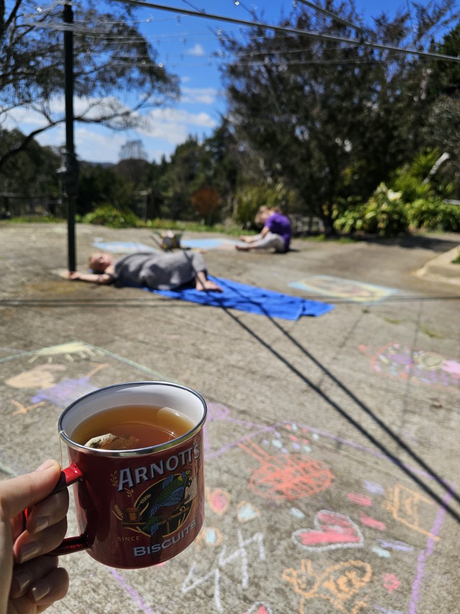 A hand holding a cup of tea, with kids playing with chalk under a hills hoist clothes line in the background.