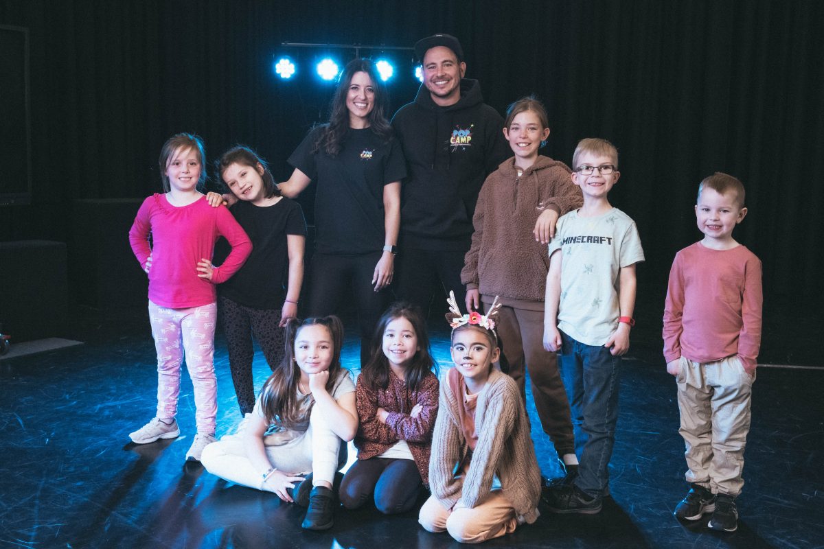 Megan and Michael Simjanovski stand on stage surrounded by children smiling. 