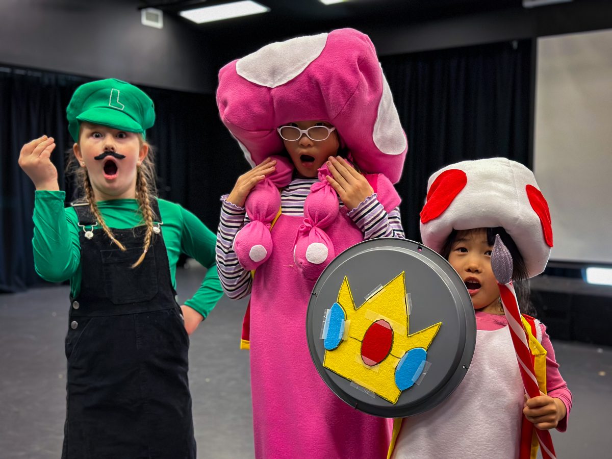 Three children stand on stage and pose, dressed in Mario Bros character costumes