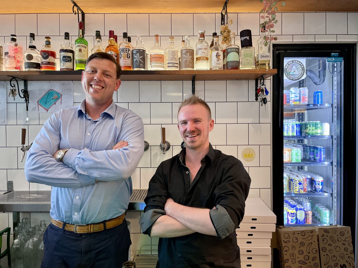 Two men stand side by side at a bar.