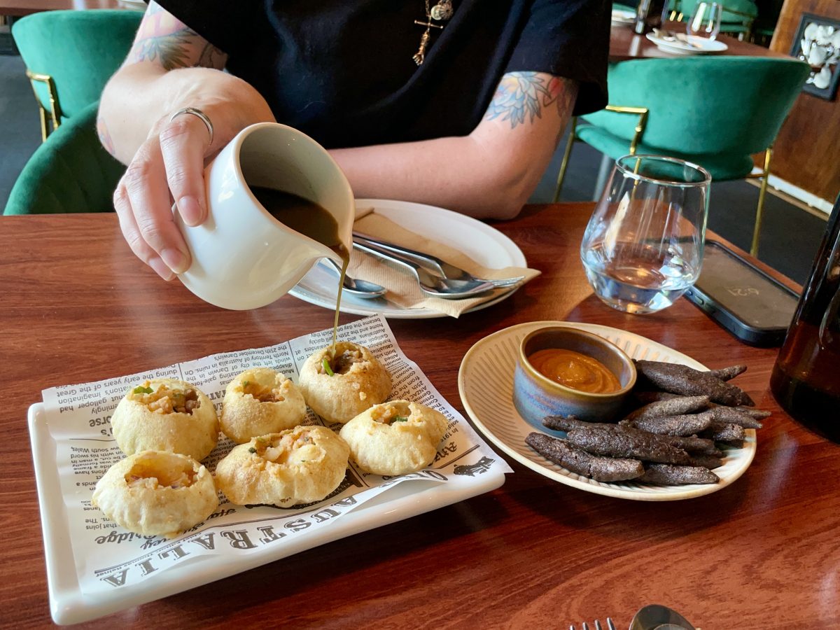 Two dishes on a table, a white hand is pouring liquid from a jug into a small circular pastry shell. 