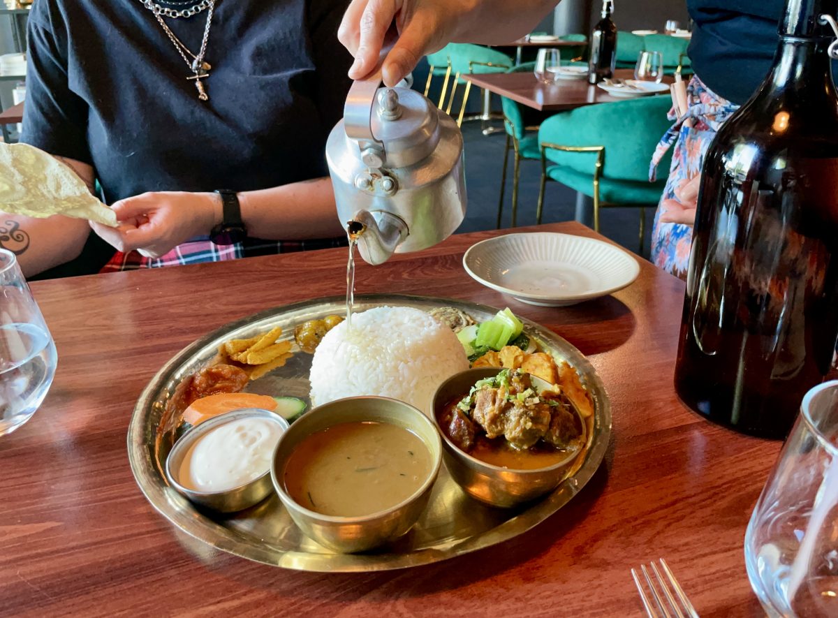 A brass platter with lots of small dishes, and a silver teapot pours ghee onto a dome of rice.