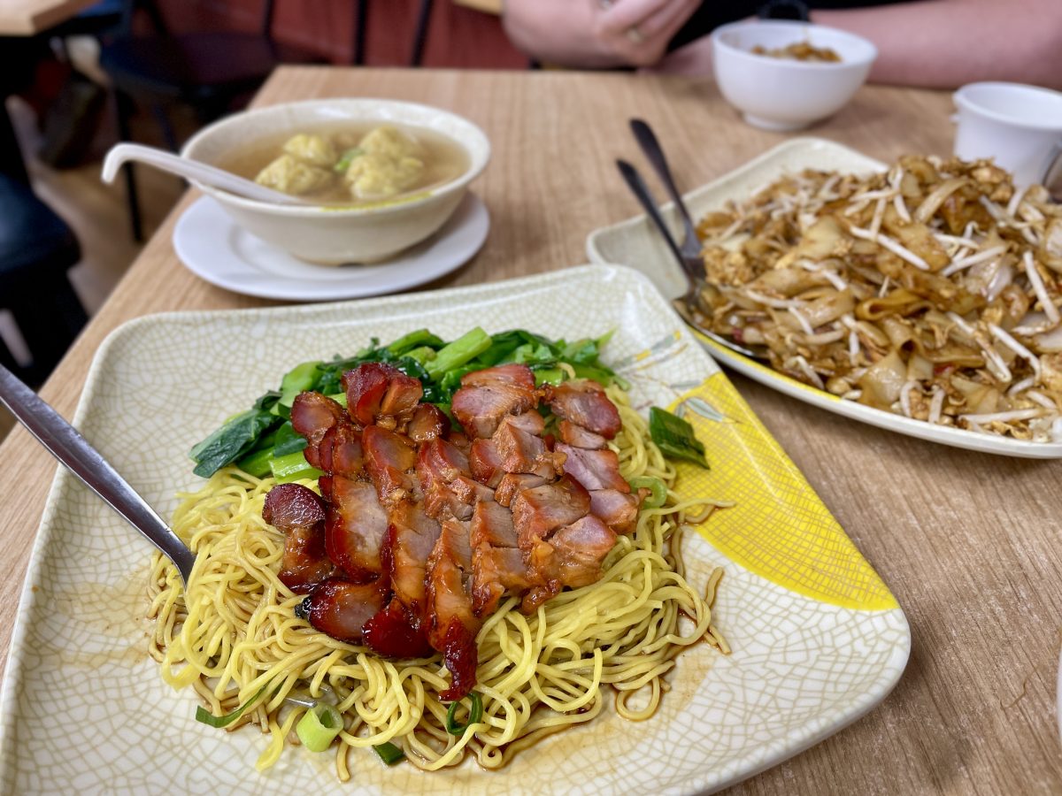 Red slices of roast pork sit on egg noodles, with greens on the side. Behind is a bowl filled with soup and four dumplings.