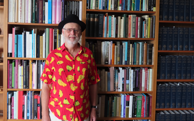 An elderly man wearing a black hat and a banana printed shirt while standing in front of a bookcase