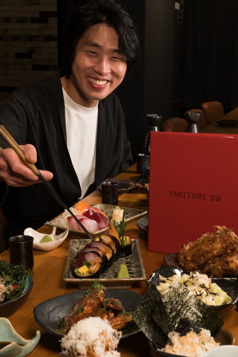 Man sits at a table laden with food and an upright menu reading Yakitori 38. He holds a piece of nigiri in chopsticks and smiles.