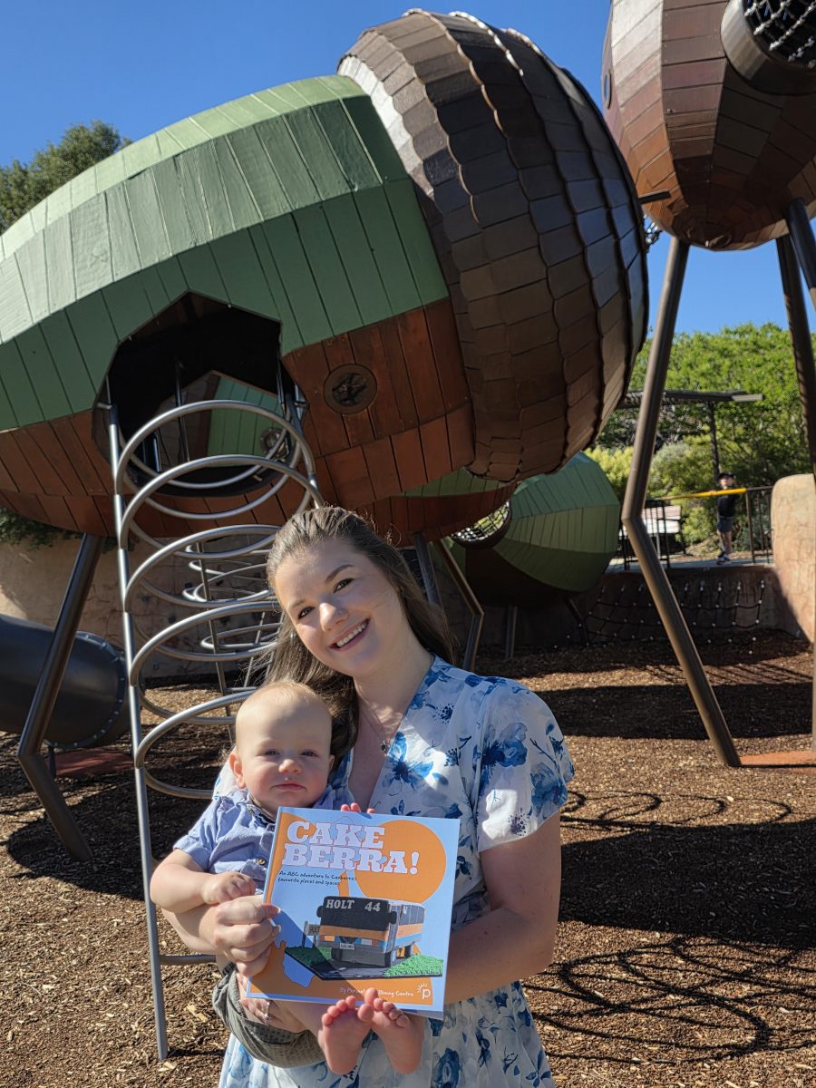 woman holding a baby and a book in front of the Arboretum's acorn playground