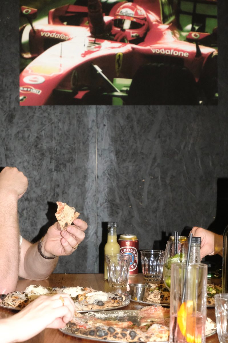A casual shot of hands holding pizzas with a poster of a race car on the wall above.