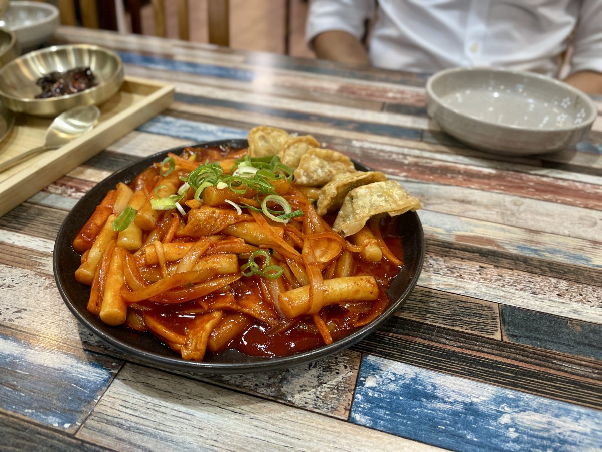 A bowl with rice cakes covered in a sticy, red, sauce and topped with crispy dumplings and spring onions.