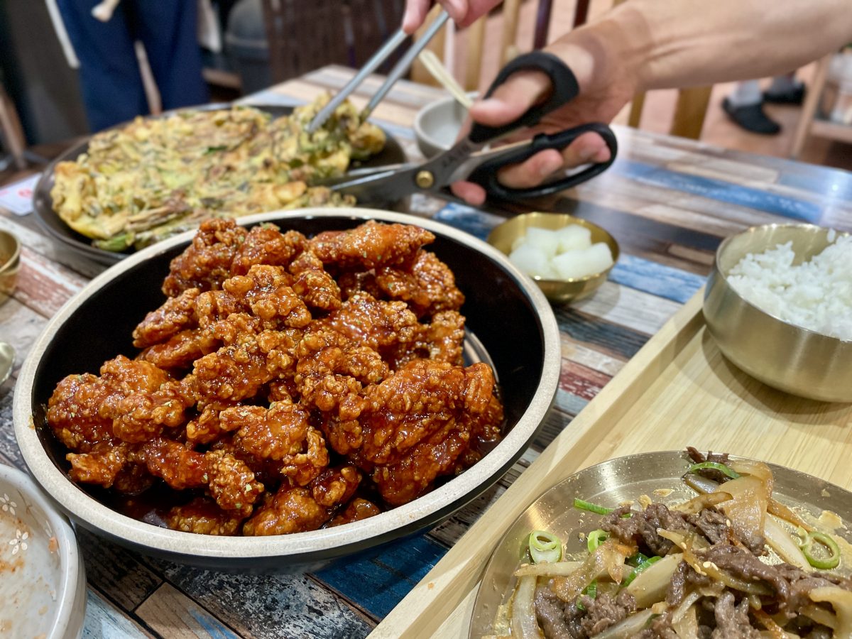 A bowl of fried chicken covered in a sticky red sauce, in the background hands cut into a savoury pancake with a pair of scissors.