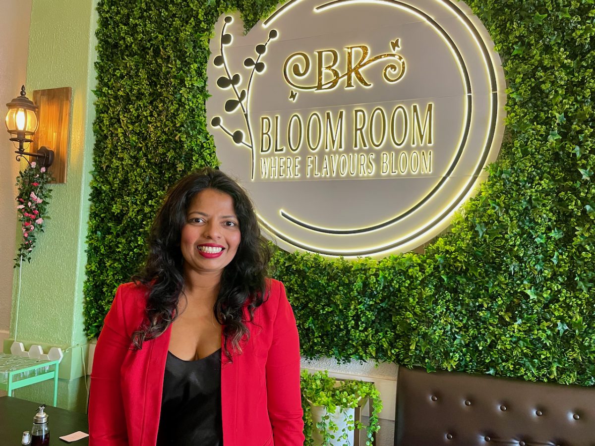 A Brown woman in red jacket stands in front of a wall covered in faux plants and a sign reading "CBR Bloom Room, Where flavours bloom"
