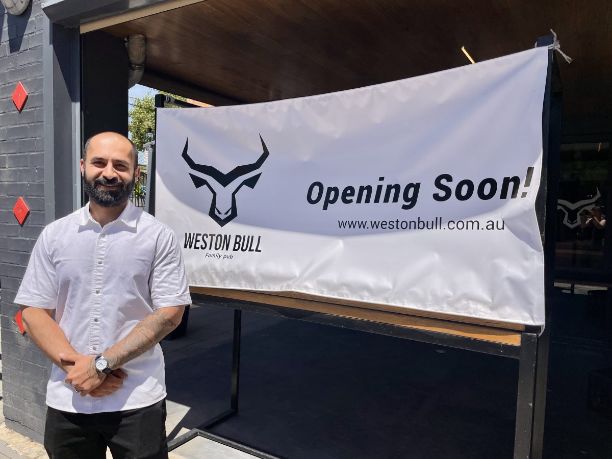 A man wearing a white shirt stands in front of a sign which reads "Opening Soon! Weston Bull family pub" with a bull horn logo.