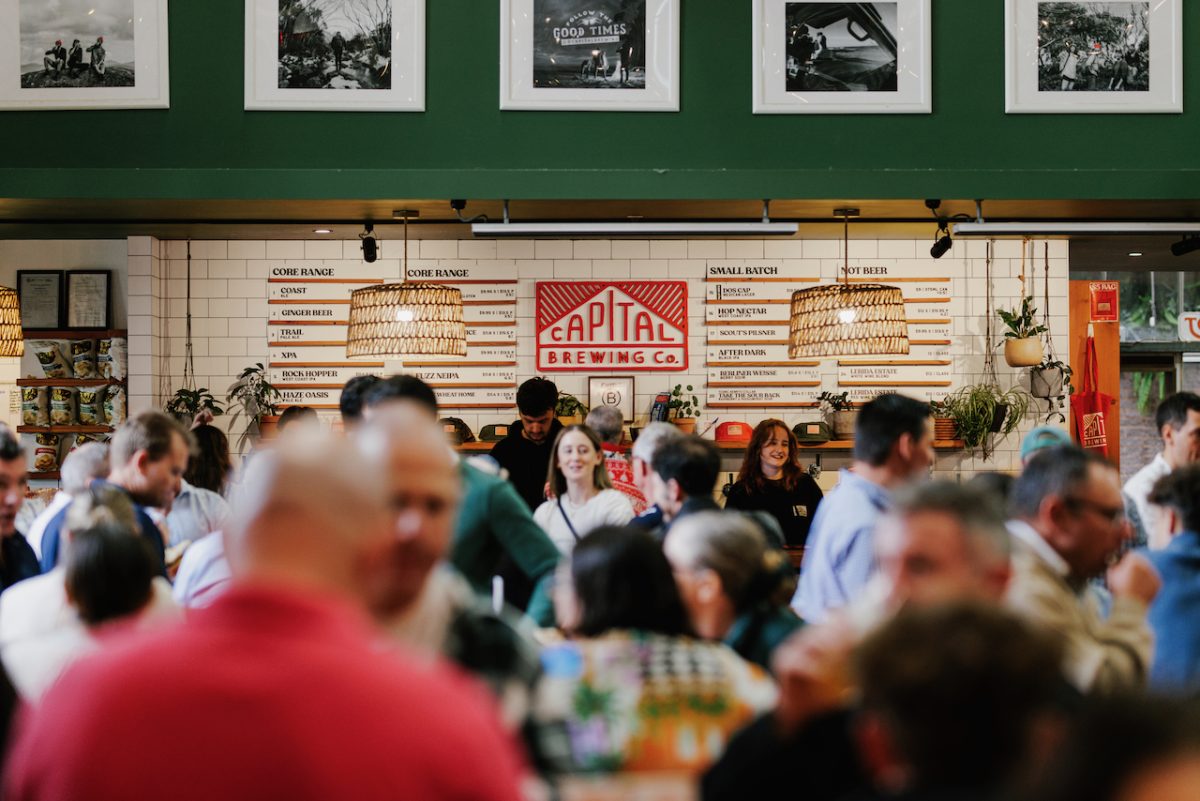 In the foreground, slightly blurred people in a crowded room. In focus behind is the bar of Capital Brewing's brew pub with their logo in the centre.