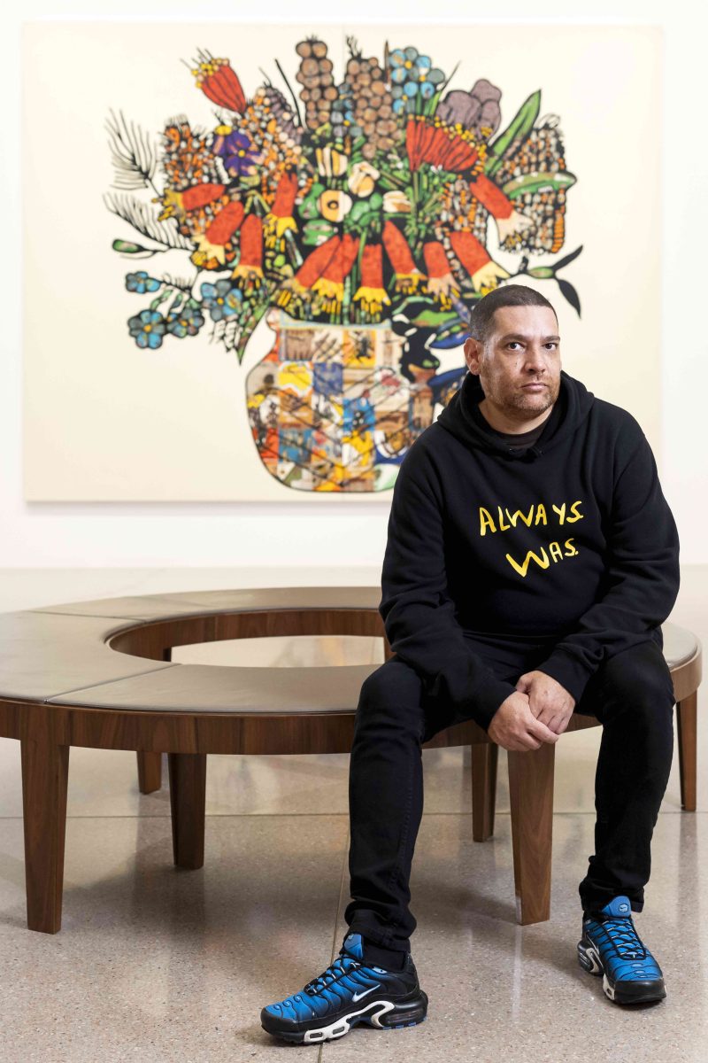 Aboriginal artist seated in front of a large flowerpiece painting