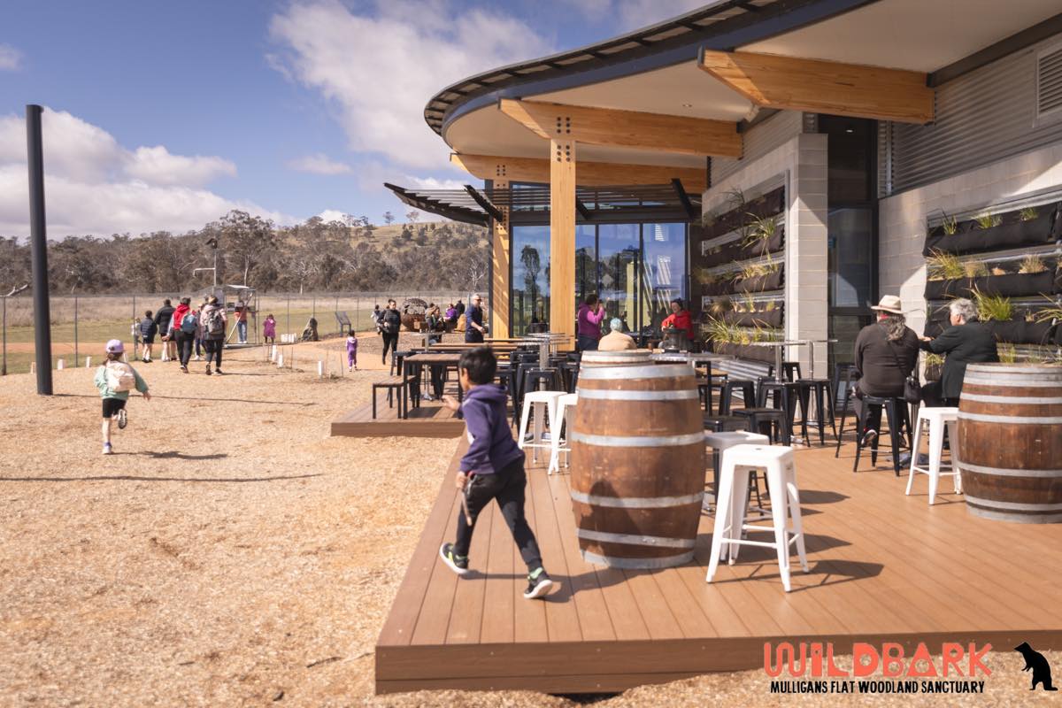 A child is running from a wooden deck towards an outdoor play space. There are cafe tables and chairs and kids playing.