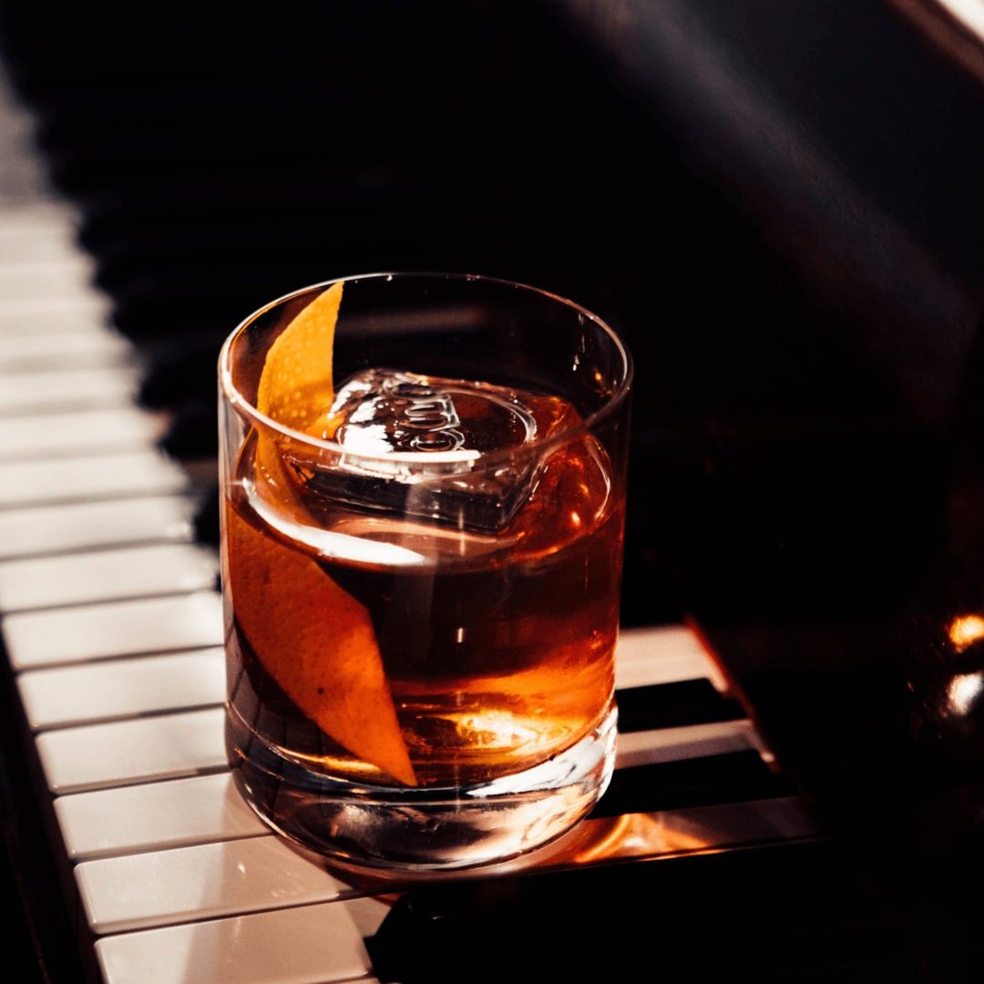 A cocktail in a tumbler with an orange peel garnish sits on a piano.