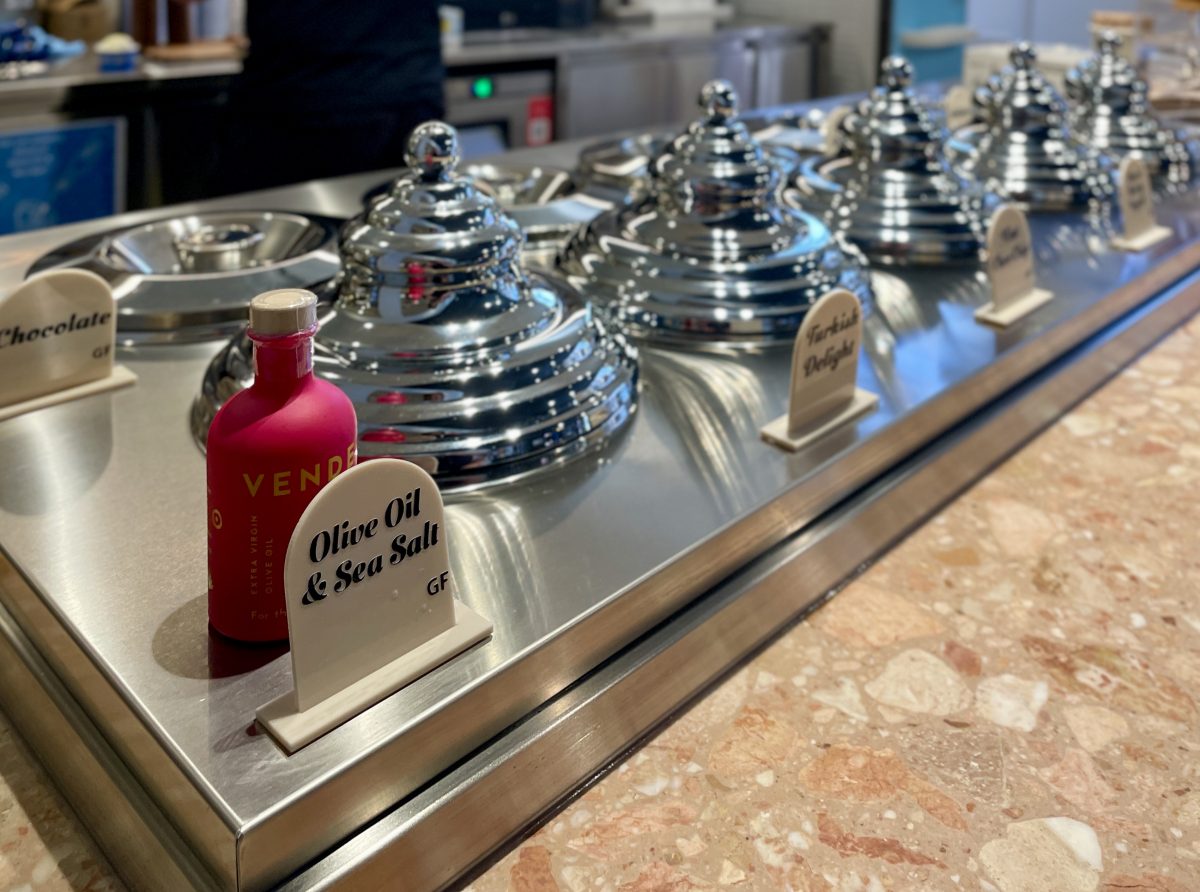 A pink bottle of olive oil behind a small sign reading 'Olive Oil & Sea Salt' on a bench with ornate silver lids covering tubs of gelati.