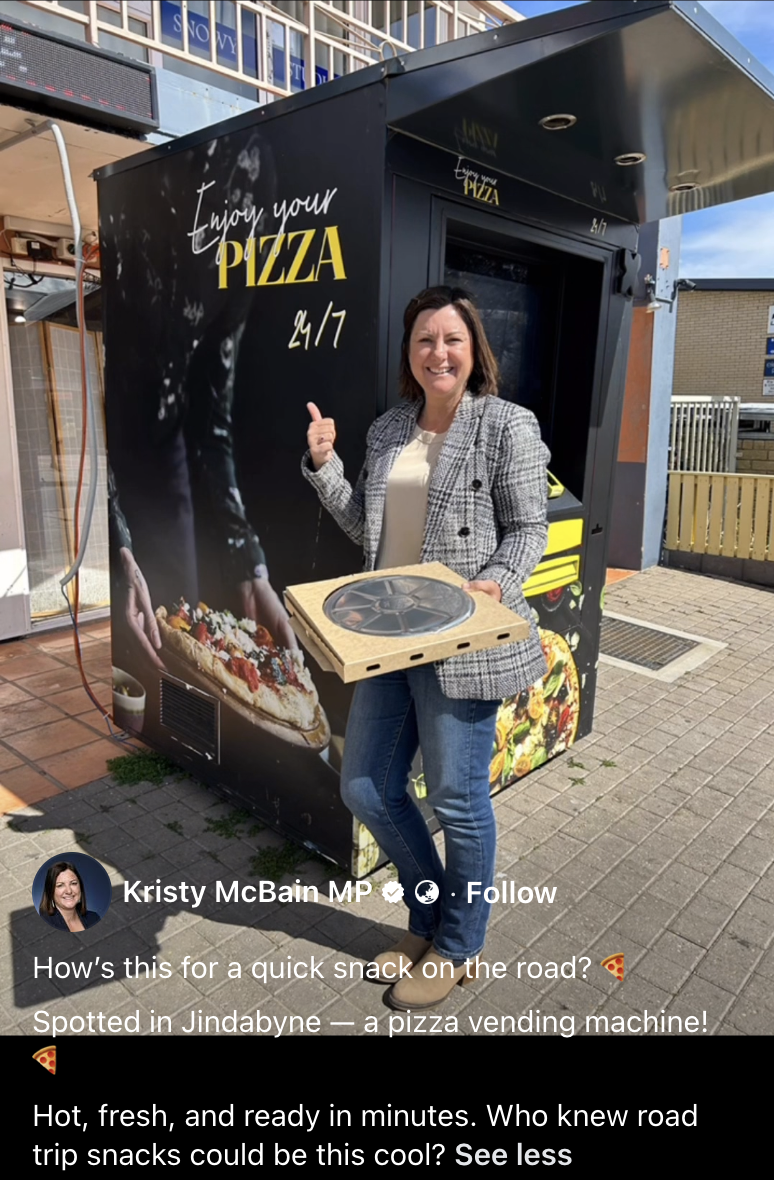 A woman wearing a grey blazer holds a pizza box with round, metal circle in the centre of the lid. She is standing in front of a black vending machine which reads "pizza 24/7" and giving a thumbs up to the camera. 
