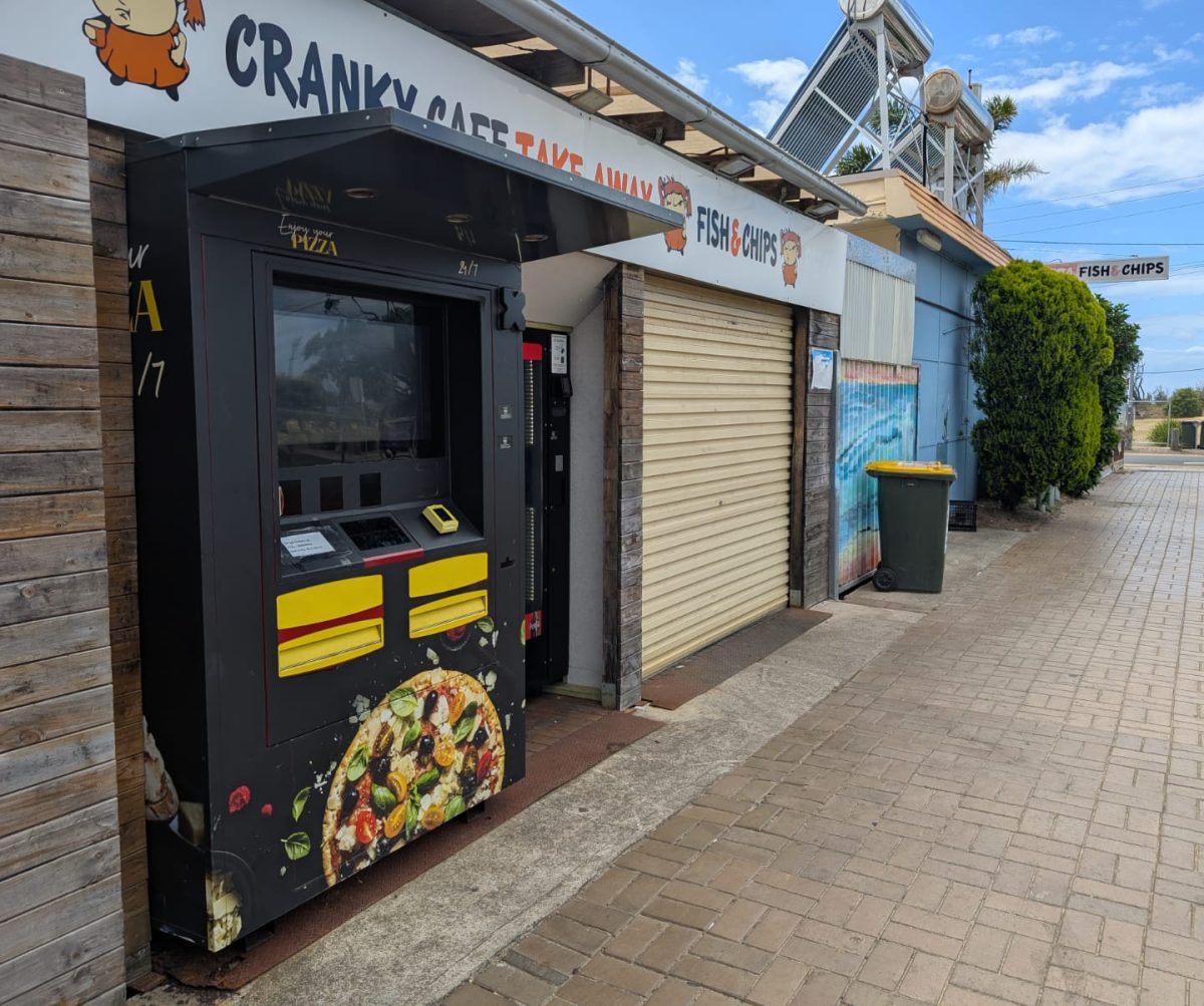 A black vending machine with image of pizza, next to a regular softdrink vending machine, under a sign reading 'cranky cafe take away fish & chips'.