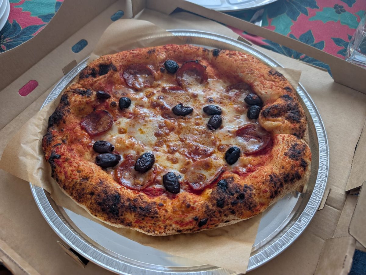 A tasty looking salami and olive pizza on a foil tray, in a cardboard pizza box.