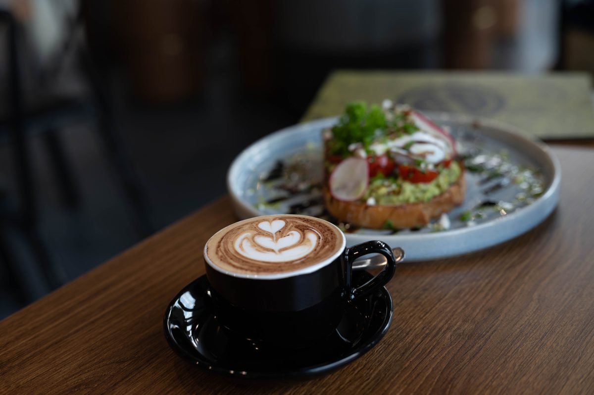 A cappucino sits on a table in front of a plate of food.