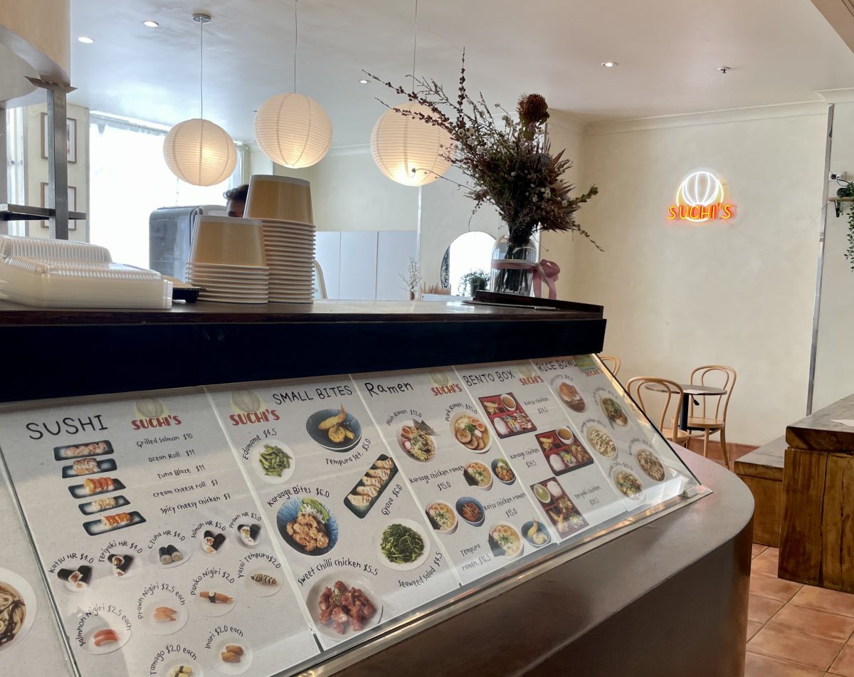 A photo of the menu at Suchi's with the cafe and neon sign in the background.