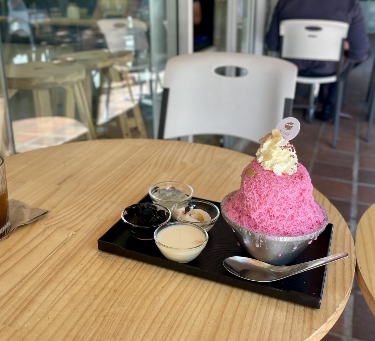A bright pink pile of shaved ice sits in a metal bowl on a tray. The ice is topped with whipped cream and almond flakes. There are bowls of additional toppings: condensed milk, black jelly squares, lychee, and translucent pearls. 